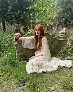 a woman in white dress sitting on grass next to stone wall and holding a red object