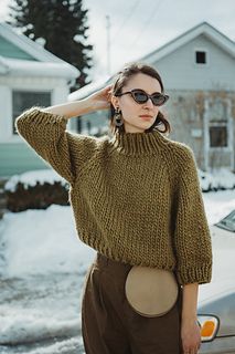 a woman standing in front of a car wearing a green sweater and brown pants with her hands on her head