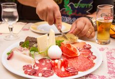 a person cutting up meat and cheese on a white plate with glasses of beer in the background