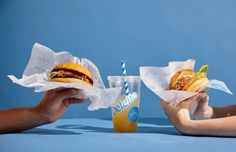 two hands holding sandwiches and a drink on a blue table with a blue wall in the background