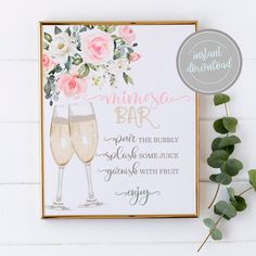 two champagne flutes with pink flowers and greenery on the side next to a framed sign