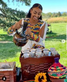 a woman is pouring tea into cups at an outdoor picnic table with other items on the grass