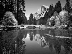 black and white photograph of a man standing on a bridge over a river with mountains in the background