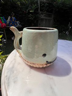 a ceramic fish mug sitting on top of a white table in front of some bushes