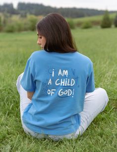 a woman sitting in the grass wearing a blue shirt that says i am a child of god