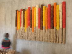 a woman walking past a wall hanging made out of yarn and wood sticks with an orange, yellow, red, and brown stripe design on it