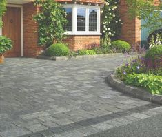 a brick driveway in front of a house with flowers and shrubs around it, along with a potted plant on the side