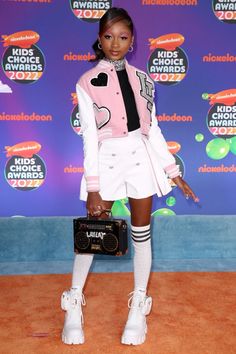 a woman in white shorts and pink jacket holding a black bag on an orange carpet