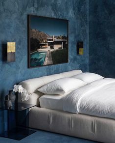 a bed with white sheets and pillows in a blue room next to a painting on the wall