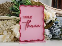 there is a pink sign that says table three on it and flowers in the background