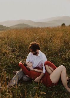 a man and woman sitting on the ground in tall grass with mountains in the background