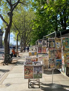 the sidewalk is lined with posters and other art work on display in front of trees