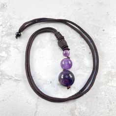 Empowerment Necklace. Lift us your style with the vibrant allure of this Amethyst pendant necklace. This pendant hangs from a brown nylon adjustable rope. Its warm, rich tones effortlessly compliment the purple amethyst pendant and adds a touch of bold sophistication to your style. This crystal necklace matches perfectly with sweaters and winter gear or even the approaching spring outfits. As a gift, its striking beauty and symbolic strength make it a thoughtful and empowering gesture. Embracing the wearer with courage and passion, this amethyst pendant necklace not only enhances your style but also serves as a meaningful addition to one's personal collection, a reminder of resilience and vitality throughout the joyous season and beyond. Adjustable Teardrop Pendant Crystal Necklace With Gemstone, Adjustable Teardrop Gemstone Crystal Necklace, Adjustable Pendant Necklace For Meditation, Purple Amulet Necklaces As Gifts, Amulet Style Necklace With Adjustable Cord For Meditation, Spiritual Amethyst Round Pendant Necklace, Amethyst Crystal Healing Necklace With Round Pendant, Amethyst Crystal Necklace For Healing, Adjustable Amulet Necklace For Meditation