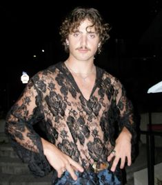 a man standing in front of stairs wearing a black lace shirt and blue denim shorts