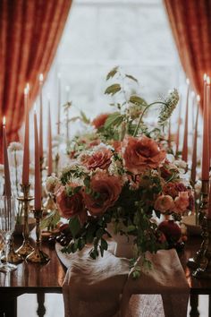 an arrangement of flowers and candles on a dining room table in front of a window