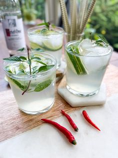 two glasses filled with ice and some mints on a cutting board next to red chili peppers