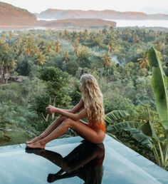 a woman sitting on the edge of a swimming pool looking out over trees and mountains