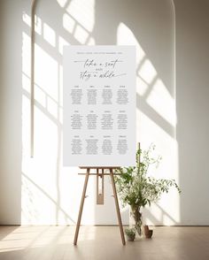 an easel with a wedding seating chart on it in front of a window and potted plant