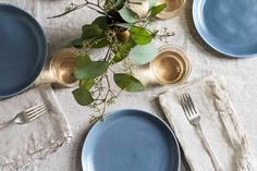 the table is set with blue plates, silverware and greenery in vases