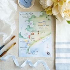 a watercolor map is on the table next to some blue and white towels, pens, and flowers