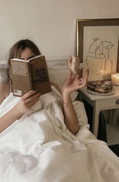 a woman laying in bed reading a book and holding a doughnut up to her face