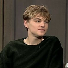 a young man sitting in front of a window wearing a black sweater and holding a silver object