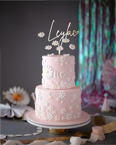 a pink cake with daisies on top and the word lyfaa spelled in gold