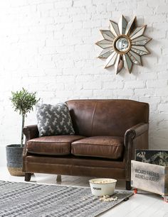 a brown leather couch sitting in front of a white brick wall next to a potted plant