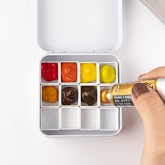 a person is holding a paintbrush and paints in an open box on a white surface