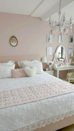 a white bed sitting under a chandelier next to a mirror and table in a room