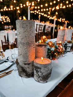 two candles are sitting on a table with white linens and lights in the background