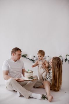 a man and woman sitting on the floor with a cake in front of them while holding a baby