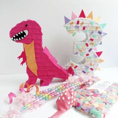 a pink dinosaur made out of paper next to a letter e and some confetti