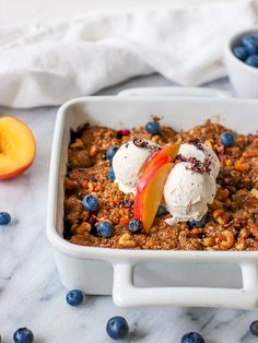 a white dish filled with blueberries, peaches and granola topped with ice cream