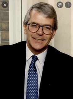 a man wearing glasses and a suit with a blue polka dot tie smiles at the camera