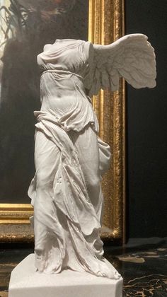 a statue of an angel standing in front of a painting on a table next to a gold frame