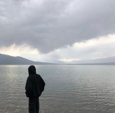 a man is standing on the shore looking out at the water and mountains in the distance