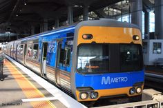 a blue and yellow metro train pulling into the station with people walking on the platform