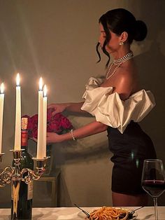 a woman standing in front of a table with food and wine on it, next to candles