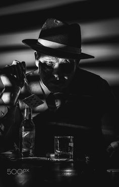 black and white photograph of a man wearing a fedora sitting at a table with liquor bottles