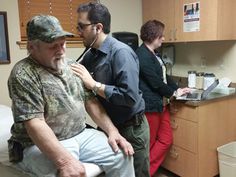 three people in a hospital room with one man getting his temperature checked by the doctor