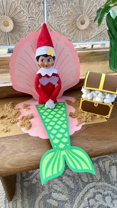 an elf is sitting on top of a table with a pink umbrella and green mermaid tail