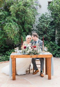 a bride and groom sitting at a table with candles on it, surrounded by greenery