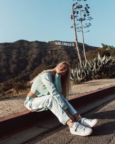 a woman sitting on the side of a road with her legs crossed and wearing white sneakers