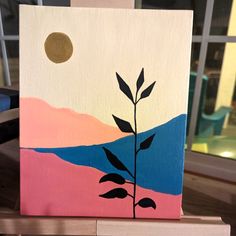 a painting on a wooden block with a plant in the foreground and a window behind it