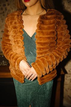 a woman wearing a brown fur jacket and green dress standing in front of a wooden table