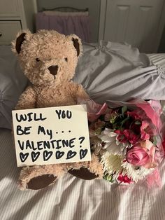 a teddy bear sitting on top of a bed next to a bouquet of flowers and a sign that says will you be my valentine?