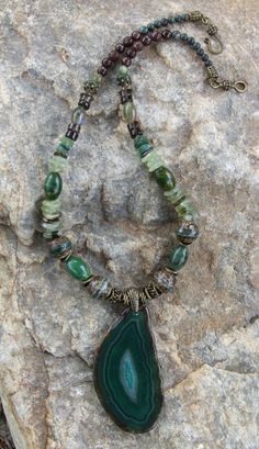 This attractive necklace is composed of many types of stones and beads:  there are 4 large tibetan line agate beads; 4 large green oval jasper gemstone beads; prehnite large chip/disc gemstone beads; fancy jasper beads; serpentine jasper beads; brown jade beads; Smokey Quartz beads; along with bronze pewter spacer beads and a hook claps, all of which are lead and nickel free. The pendant is 3 inches long and 1 1/2 inches wide, with crystals in the middle, framed by dark and light swirls of green and a few brown swirl lines.  The necklace is 20 1/4 inches long. Luxury Agate Beaded Necklace With Natural Stones, Agate Necklace Green, Luxury Beaded Agate Necklaces, Luxury Long Agate Necklace, Luxury Green Agate Beaded Necklaces, Bohemian Green Onyx Necklaces With Natural Stones, Bohemian Green Onyx Necklace With Natural Stones, Green Pendant Beaded Necklace With Natural Stones, Green Beaded Pendant Necklace With Natural Stones