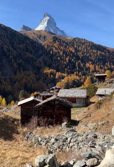 an old cabin in the mountains with a mountain behind it and trees that are changing colors