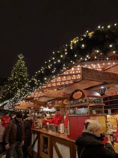 many people are standing around at the food stand with christmas lights on it and trees in the background